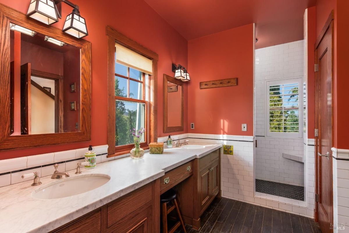 Bathroom has red walls, double vanity with wooden cabinets, a walk-in shower, and two windows.