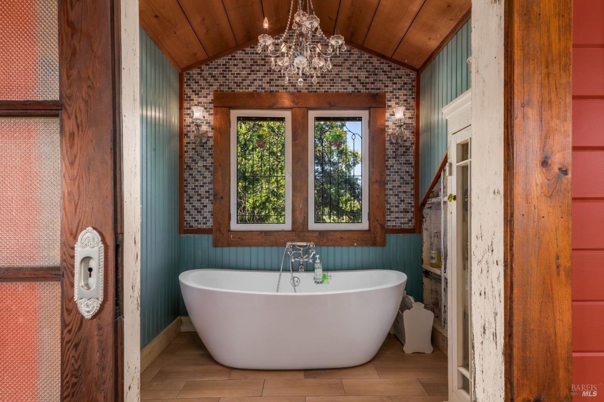 Bathroom includes soaking tub, chandelier, two stained glass windows, and wood-paneled walls.