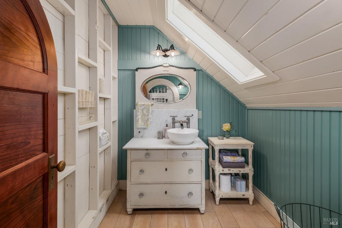 Bathroom has blue paneled walls, a dormer window, a repurposed dresser as a vanity, and built-in shelving.