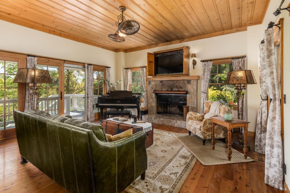 Living room with stone fireplace, grand piano, and large windows opening to a deck.