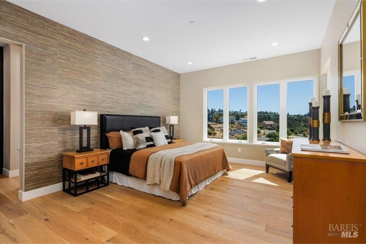 Master bedroom combines a warm accent wall with large windows offering natural light and a cozy seating area.