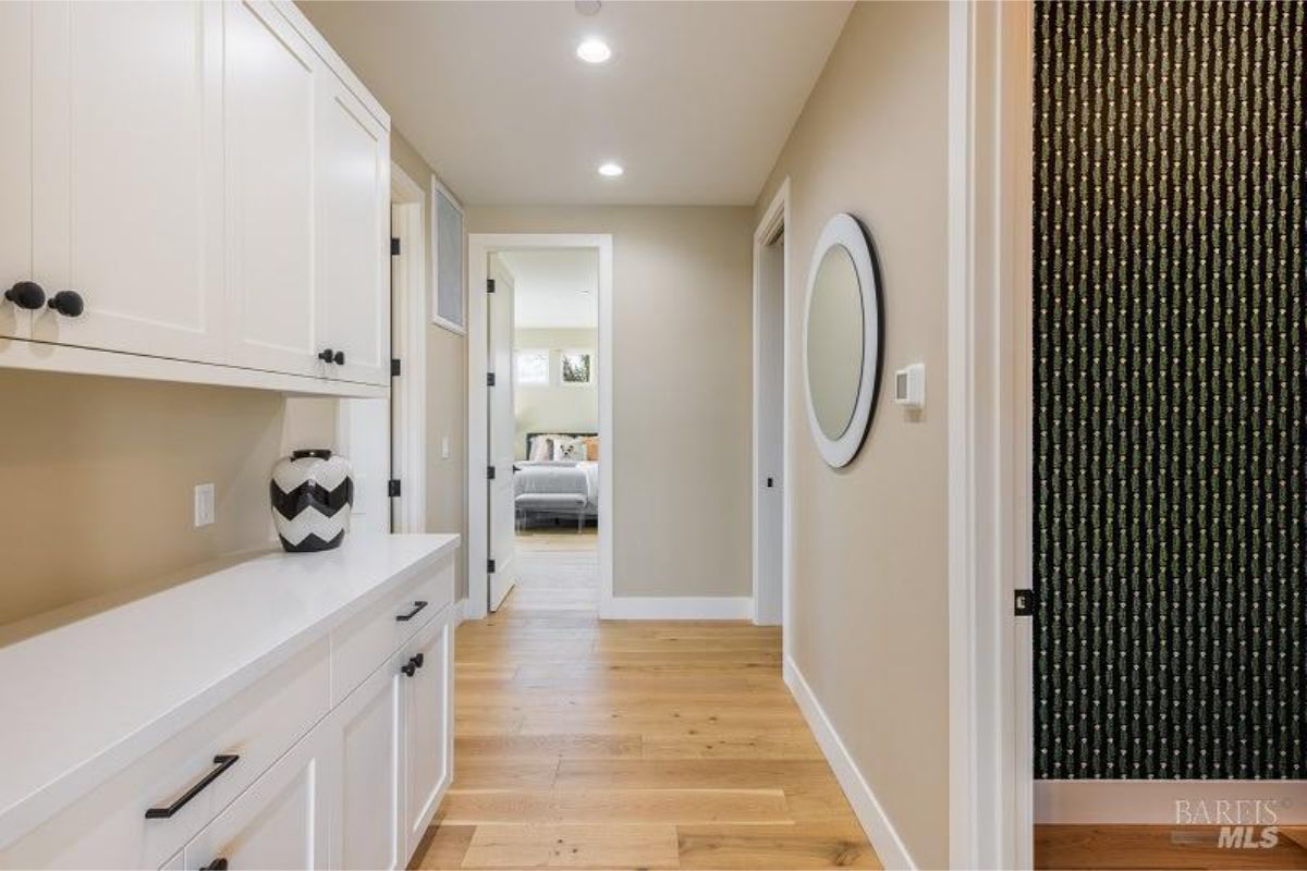 Hallway has wood floors, white built-in cabinets, a round mirror, and wallpaper.