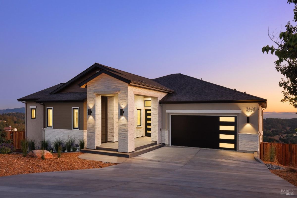 Modern, one-story home with a large garage door, a covered porch, and a stone facade at dusk.