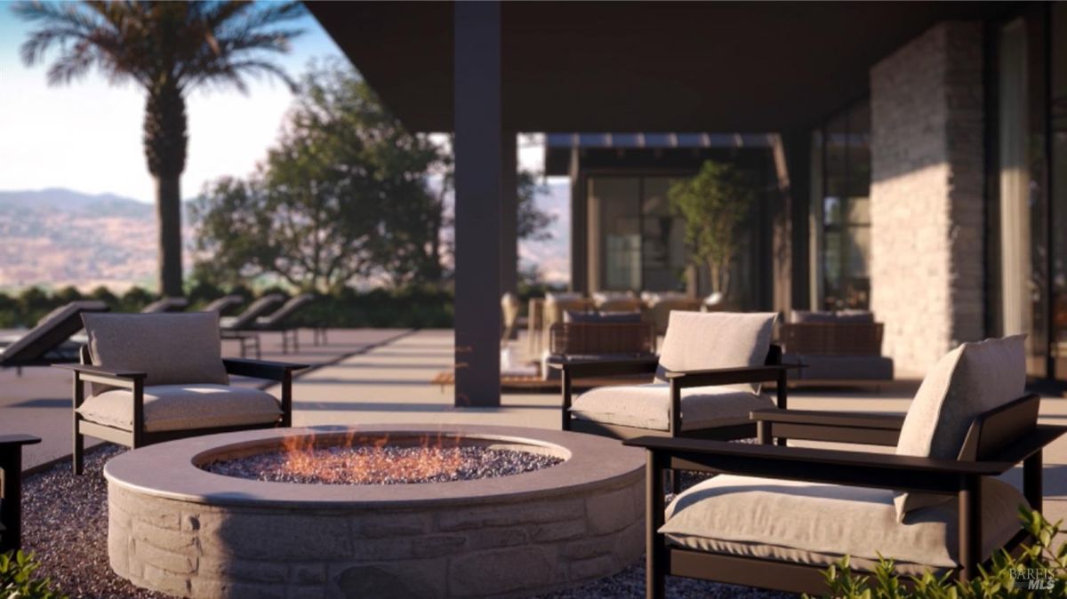 Shows a stone firepit surrounded by cushioned outdoor chairs on a patio with views of landscaped greenery and hills.