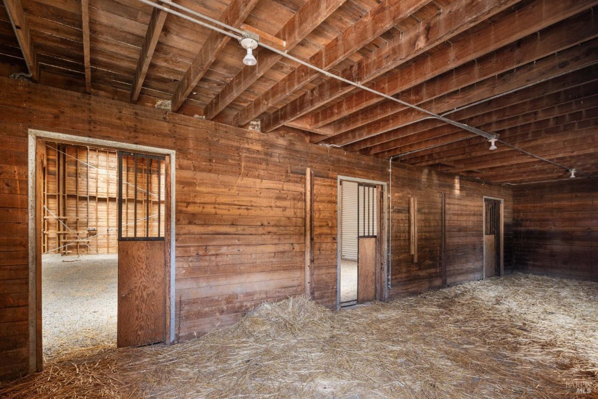 An interior view of a rustic barn with wooden walls, hay scattered on the ground, and open stall doors.