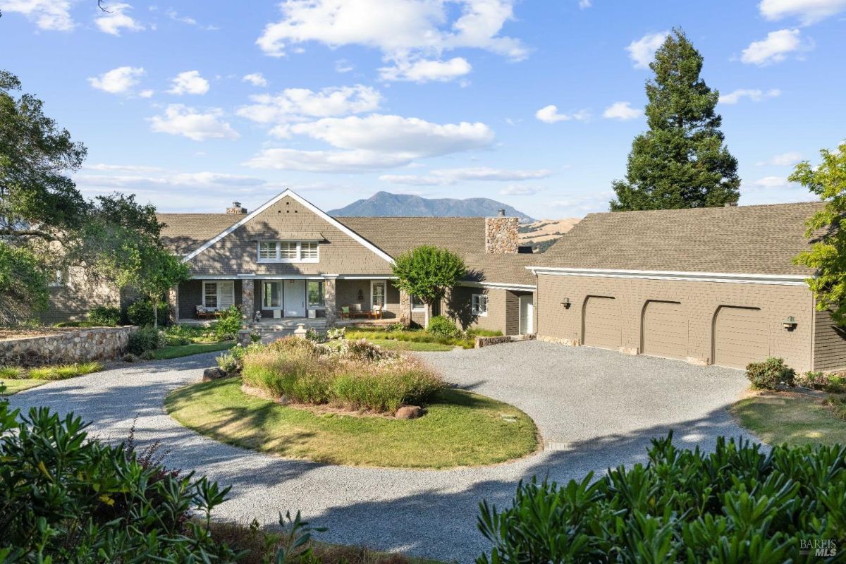 Charming country-style home with a circular driveway and a three-car garage, set against a mountainous backdrop.