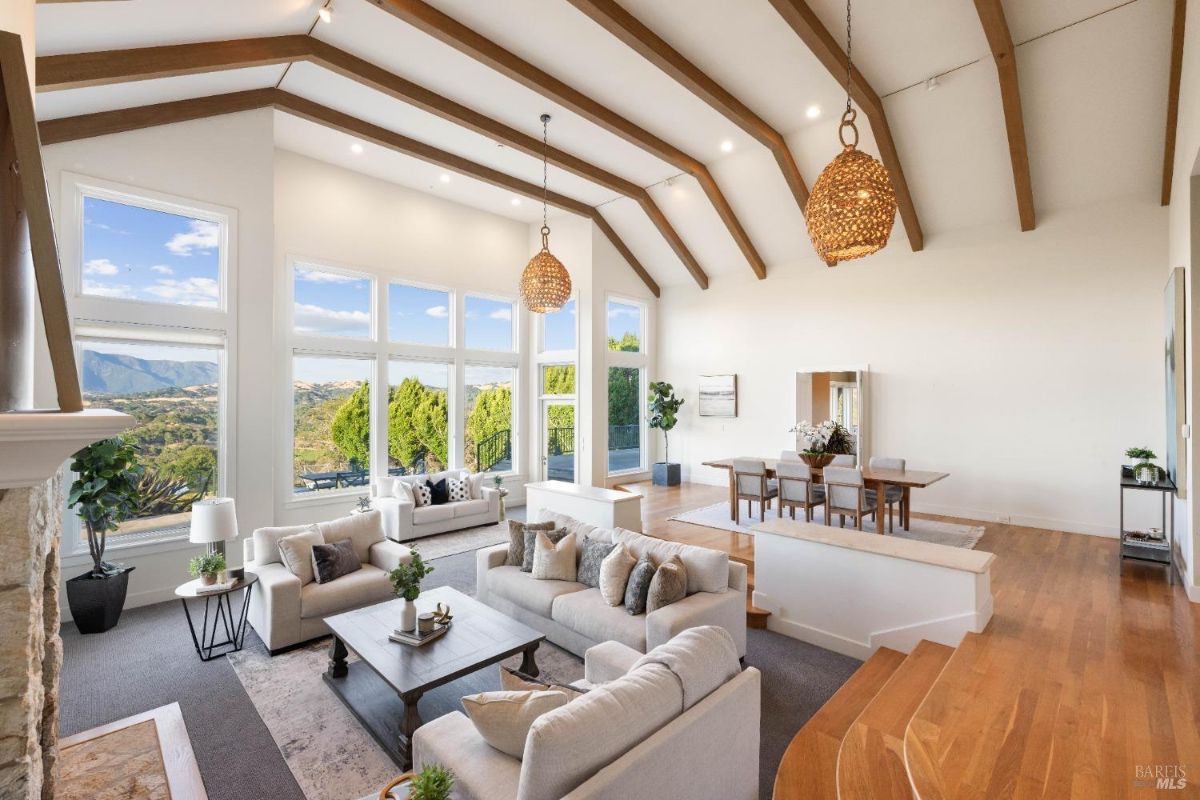 A spacious and elegant living room with large windows offering panoramic views of rolling hills and a bright, airy design featuring exposed beams and neutral tones. 