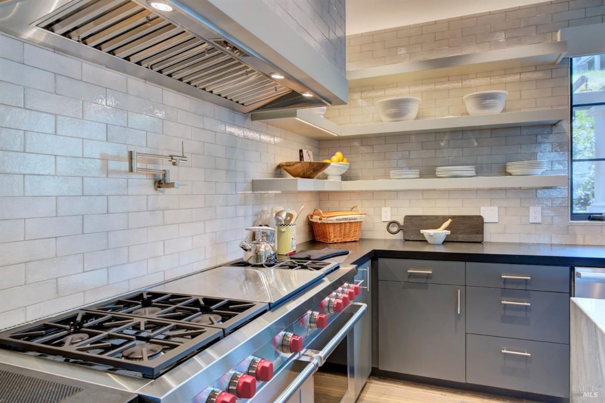 A close-up of a professional-grade stove, pot filler, and open shelves on a subway-tile backsplash.