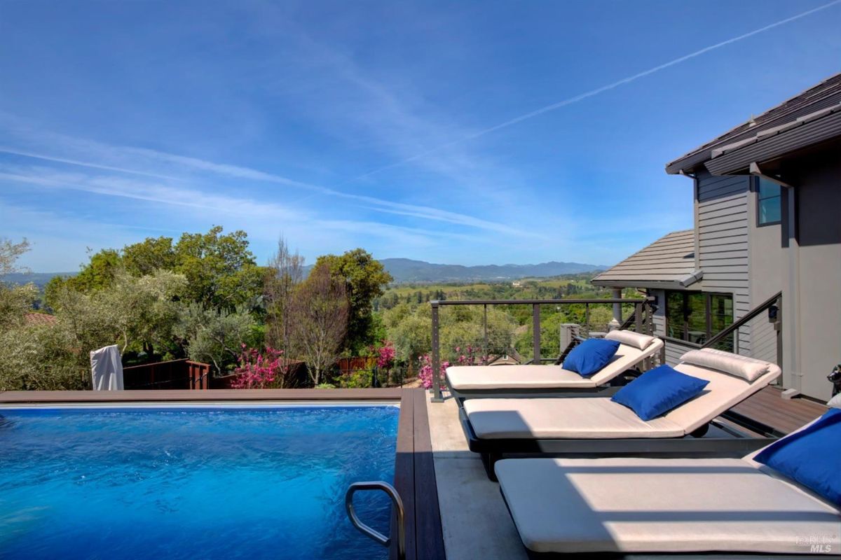 Poolside with lounge chairs with pillows on a deck overlooking the sunset and distant hills.