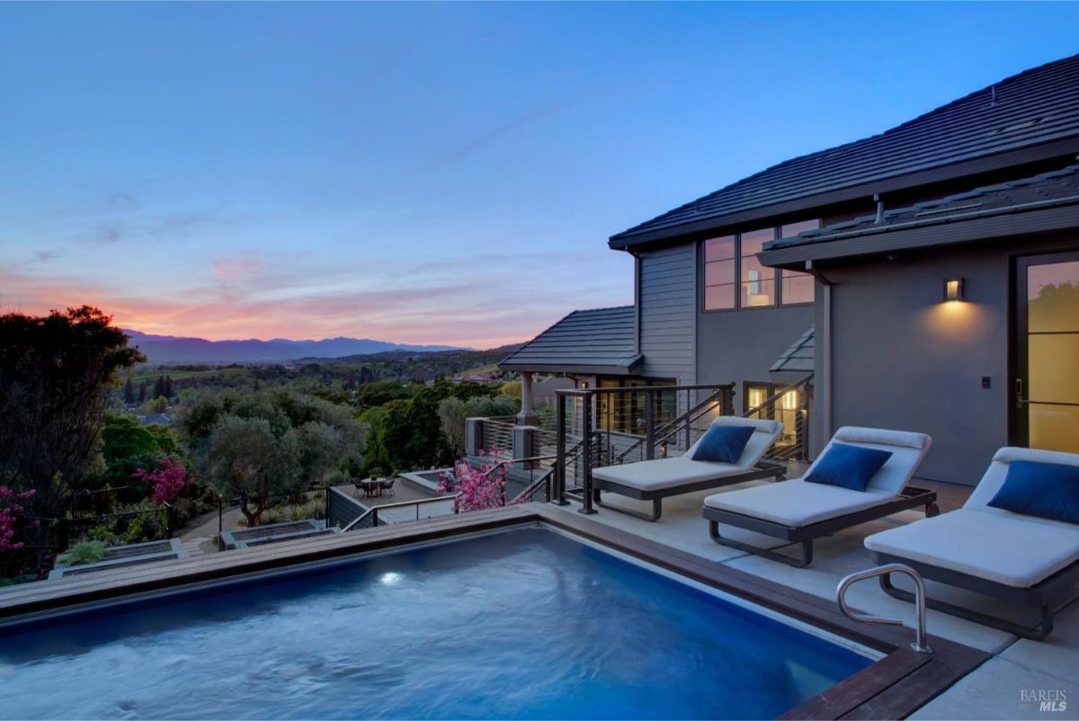 Poolside with lounge chairs with pillows on a deck overlooking the sunset and distant hills.