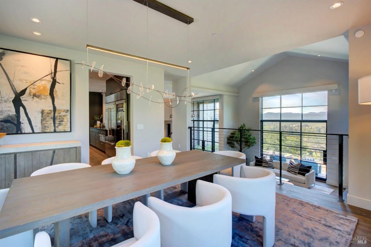 Dining area adjacent to a living space with expansive windows and a modern chandelier above the table.