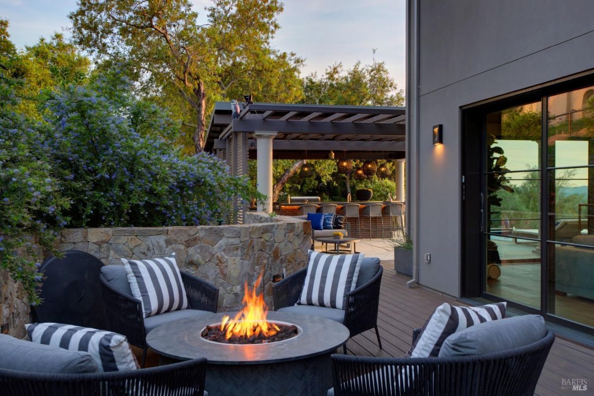 Outdoor firepit area surrounded by seating and greenery, with a pergola and bar area in the background.