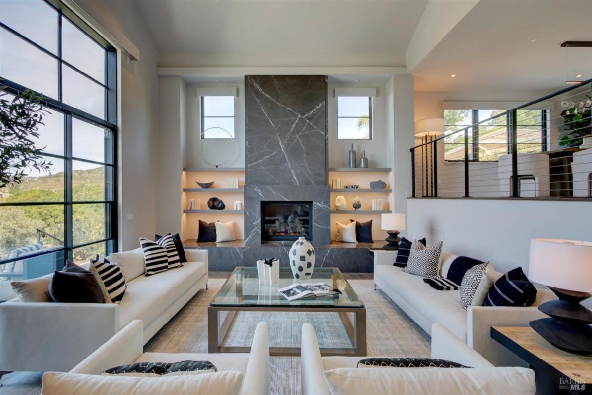 The interior living room with a central fireplace, built-in shelves, modern furniture, and large windows offering natural light.