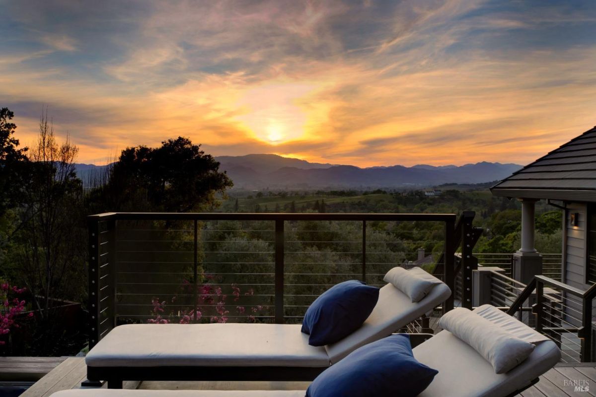 Lounge chairs on a balcony facing a scenic sunset with mountains and trees in the distance.