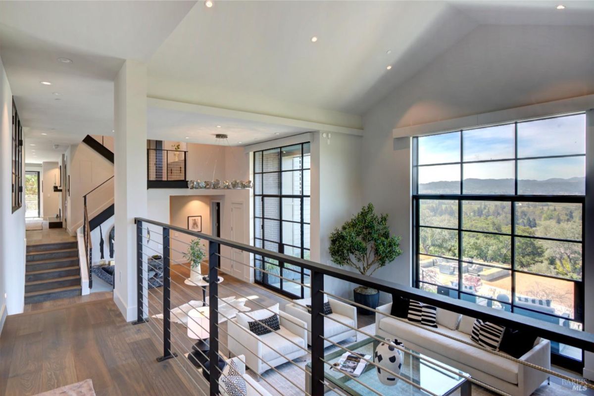 An upper-level view of the living room with large windows and a balcony-style hallway.