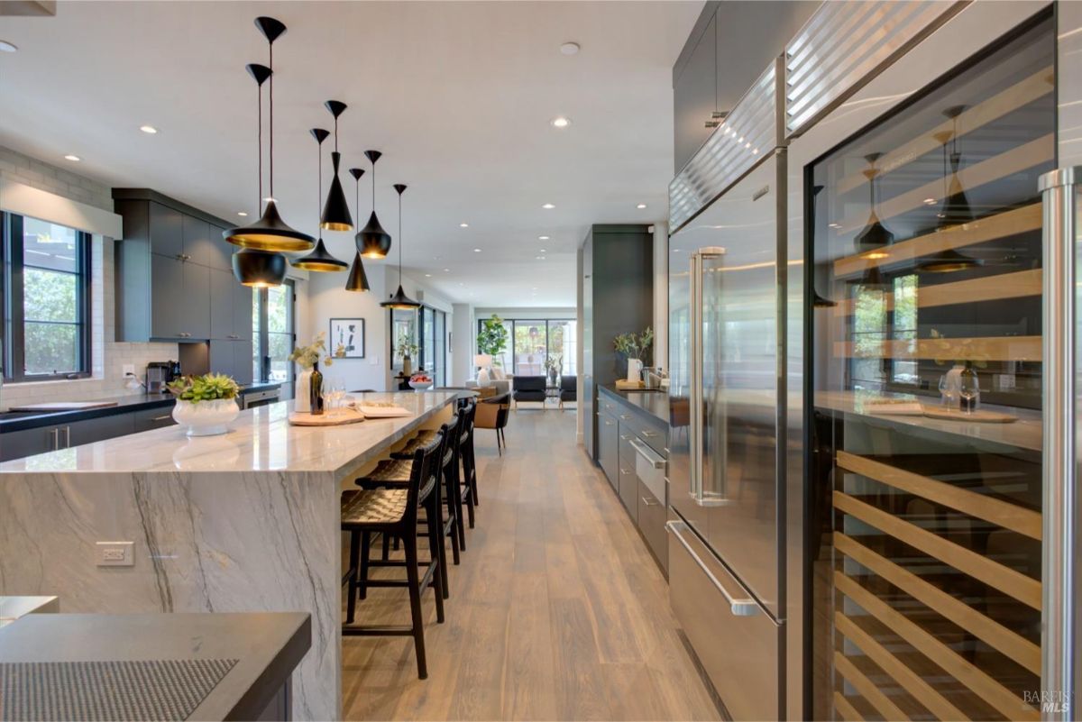 A wider view of the kitchen featuring stainless steel appliances, a wine cooler, and a long marble countertop with bar seating.