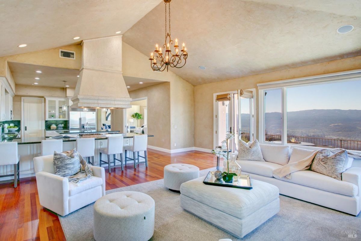 Living room with a central white sofa, armchairs, and an ottoman, featuring a chandelier and a view through sliding glass doors.