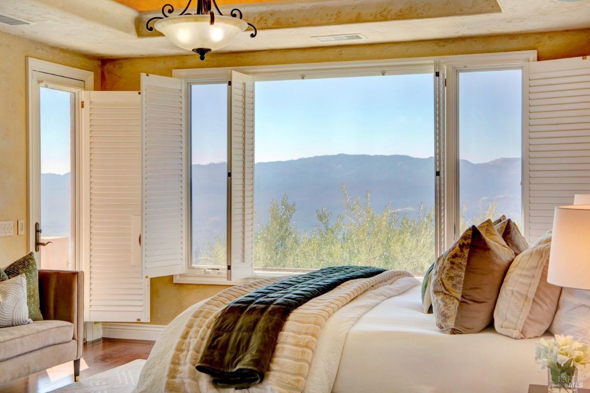 Bedroom with corner windows, white shutters, and a seating area offering views of the landscape.