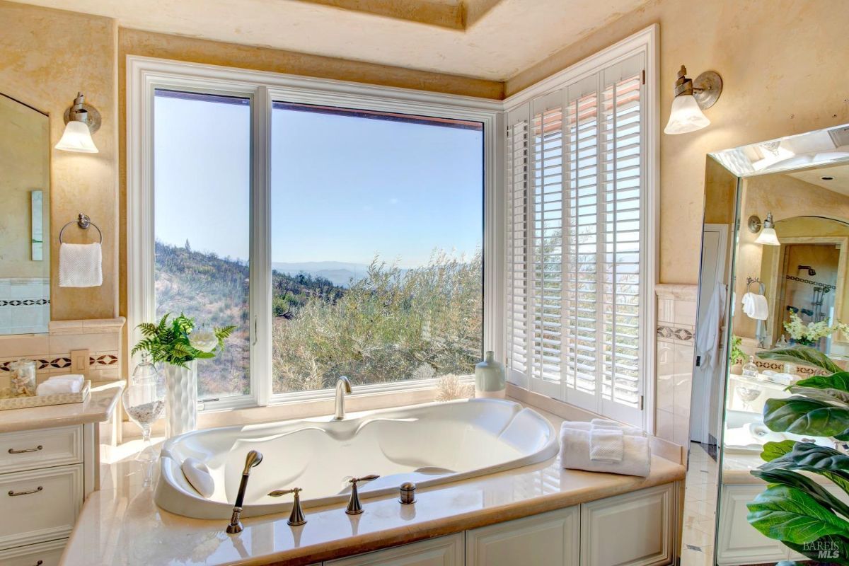 Bathroom showcases a soaking tub with a scenic view through large windows and white shutters.