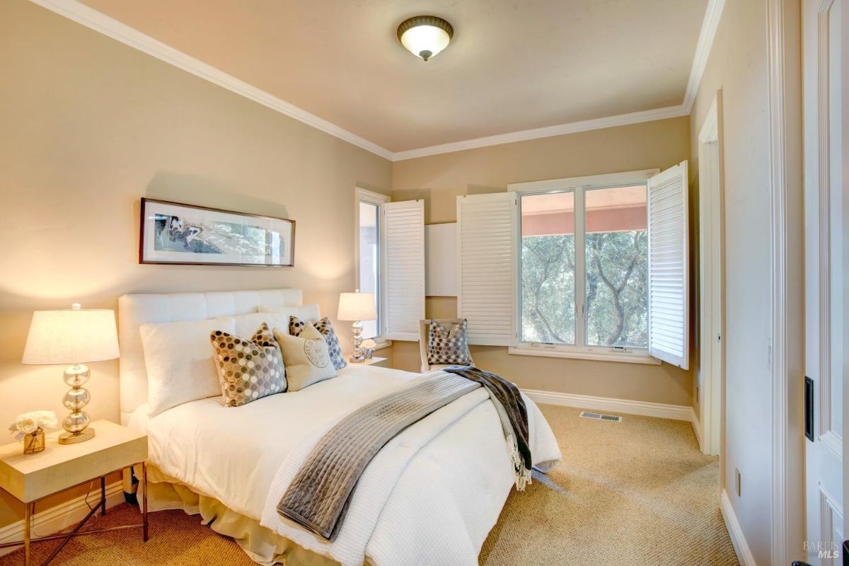 Bedroom with beige walls, white bedding, and large windows with shutters overlooking trees.