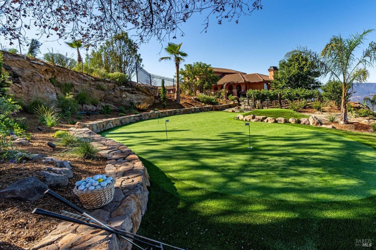 Landscaped backyard with a putting green surrounded by stone edging, palm trees, and a view of a Mediterranean-style home.