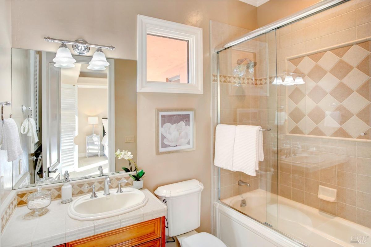 A single vanity, a framed mirror, and a glass-enclosed bathtub with beige tiles.