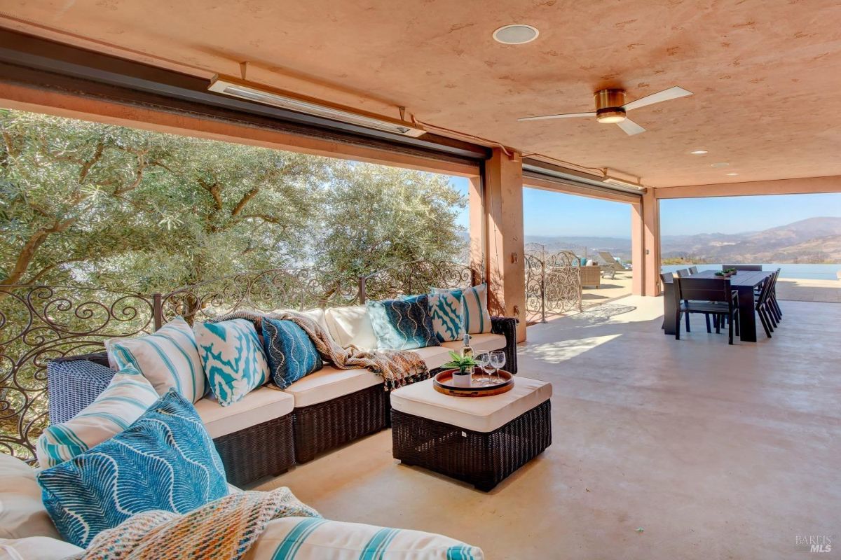 Covered patio area features a sectional sofa with patterned pillows and wrought iron railing offering partial views of the landscape.