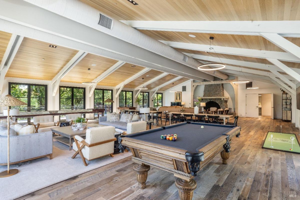 Spacious recreational area with a pool table in the foreground and a small putting green to the side. Seating areas with sofas, chairs, and coffee tables are arranged on a large rug, facing windows that provide ample natural light. A stone fireplace is located at the far end of the room, surrounded by additional tables and chairs for dining or activities.