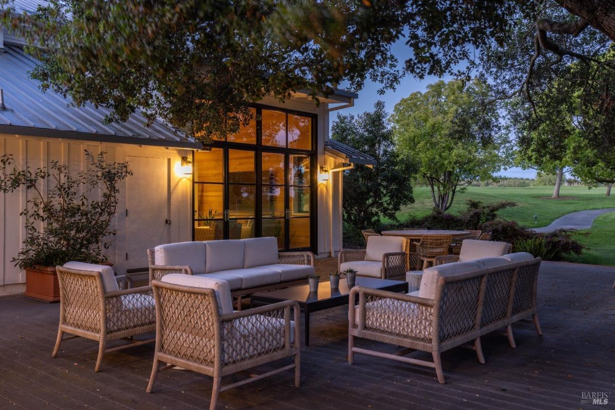 Outdoor patio area with cushioned wicker seating arranged around a rectangular coffee table. A large tree provides overhead shade, and the setting is adjacent to a building with large glass windows and doors. In the background, a green lawn and trees extend to the horizon, with a walking path curving through the landscape.