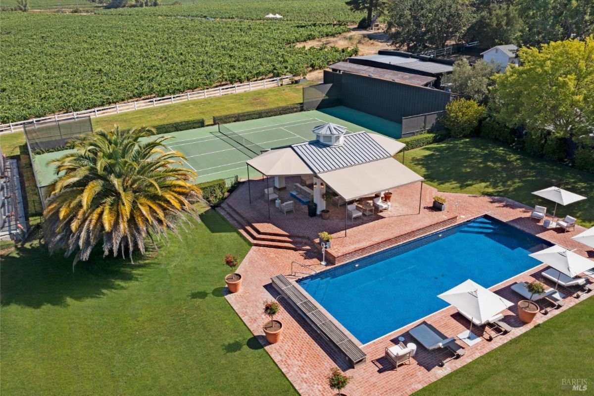 A rectangular swimming pool surrounded by a red brick deck with lounge chairs and umbrellas. Adjacent to the pool is a covered seating area with a gray metal roof, outdoor furniture, and potted plants. A tennis court with a green surface is visible in the background, alongside a large palm tree and surrounding greenery.