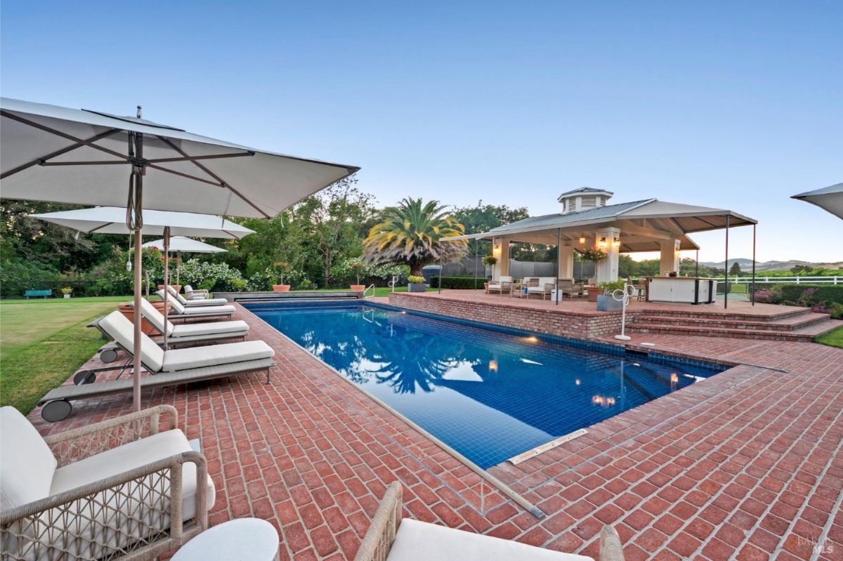 Rectangular swimming pool surrounded by a brick deck with several lounge chairs and umbrellas on one side. A covered outdoor seating and dining area is visible in the background, along with greenery and a distant view of a fence and landscape. The pool reflects the surrounding trees and umbrellas, and there are potted plants placed around the deck.