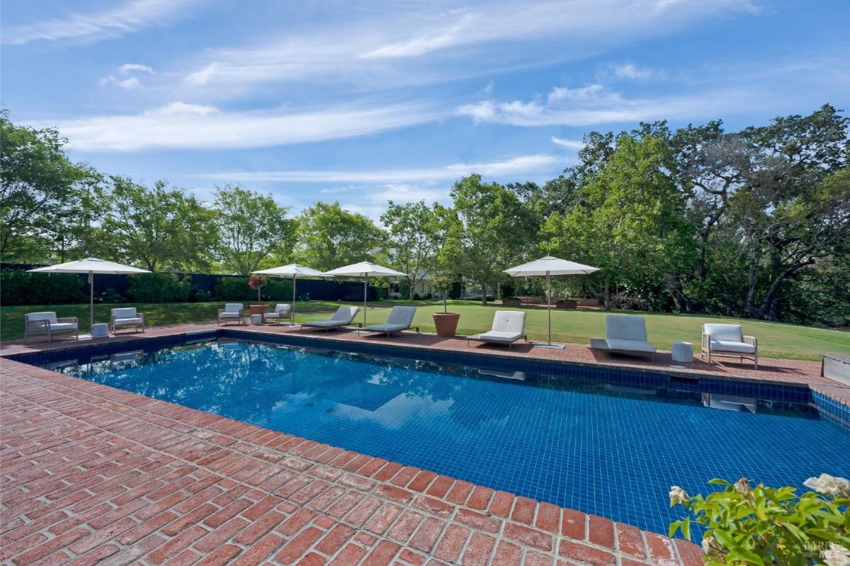 Swimming pool surrounded by red brick decking with lounge chairs and large umbrellas evenly spaced around it. Green grass and trees are visible in the background, providing a natural border to the area. The pool water reflects the sky, and potted plants are placed alongside the seating arrangements.