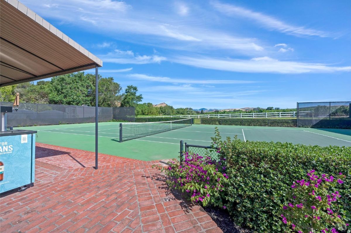 A tennis court with green playing surfaces surrounded by a black fence. In the foreground, there is a red brick-paved patio area with flowering bushes and a covered structure providing shade. The background features open fields, a white fence, trees, and distant hills under a clear blue sky.