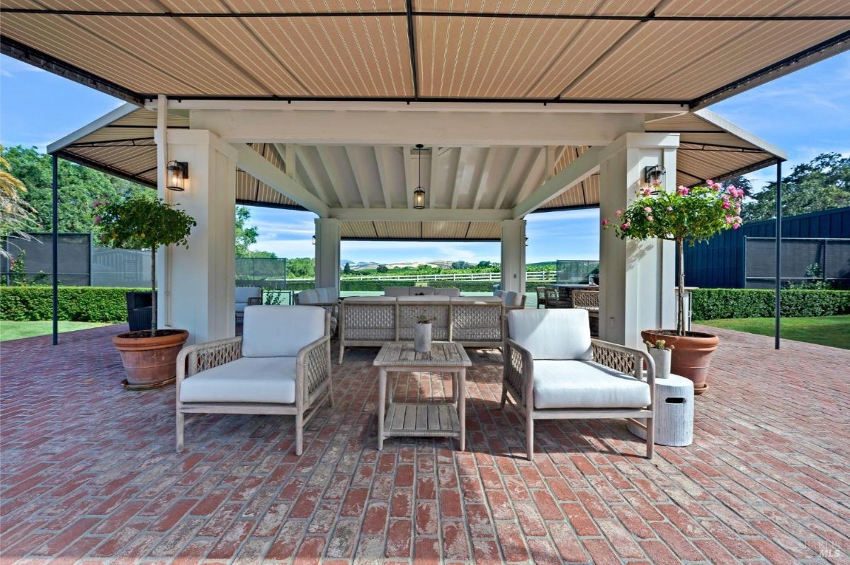 Outdoor covered seating area with a brick floor and two wicker armchairs with cushions facing a small wooden table. Potted plants with hanging flowers flank the support columns on either side of the structure. In the background, there is a view of a fence, greenery, and open fields.