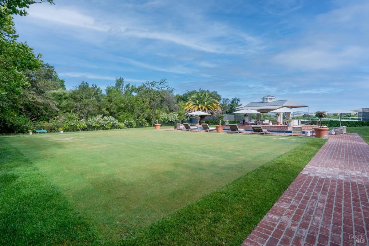 A large grassy area surrounded by trees and bushes. A red brick pathway borders the lawn and leads to a poolside area with lounge chairs, umbrellas, and potted plants. In the background, there is a covered pavilion with seating and a structure resembling a cabana near the pool.