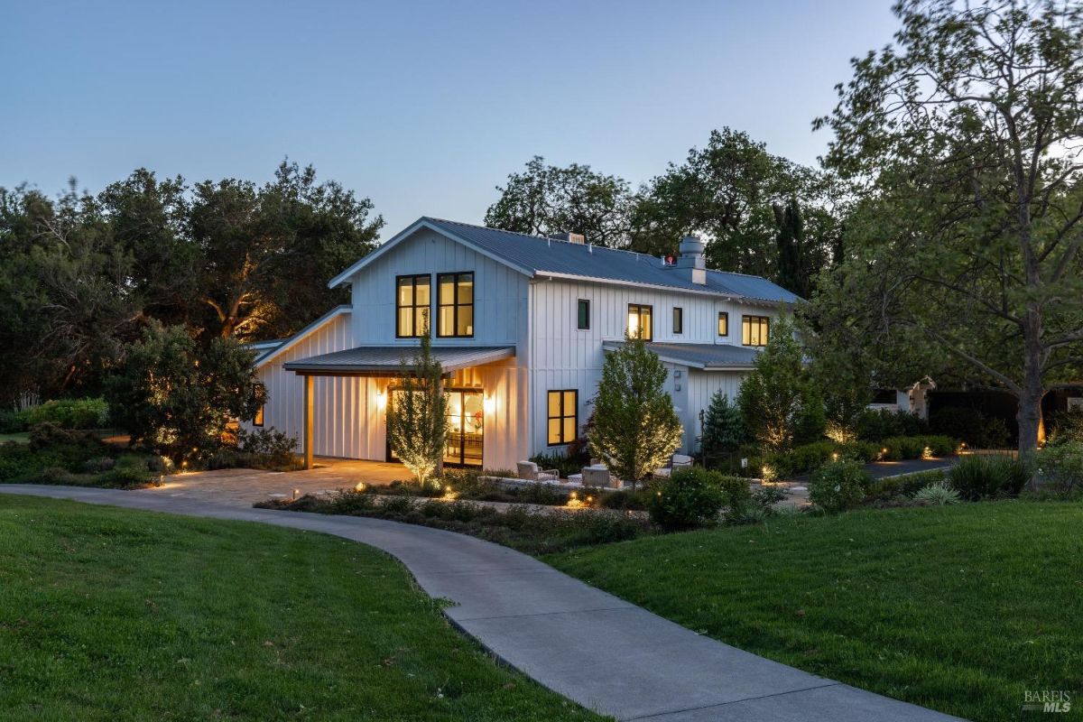 Two-story house with a modern farmhouse design, featuring white exterior walls, black-framed windows, and a metal roof. Outdoor lighting illuminates the surrounding trees, landscaping, and the pathway leading to the house. The house is surrounded by a lawn and mature trees, with a concrete walkway curving toward the entrance.