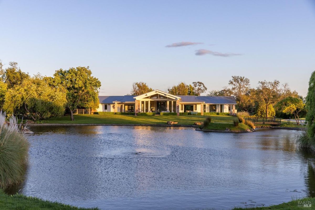 Modern single-story house with large windows, situated on a green lawn near a pond. A wooden footbridge crosses over the pond leading toward the house. Trees and shrubs surround the area, and the pond reflects the sky and part of the house.