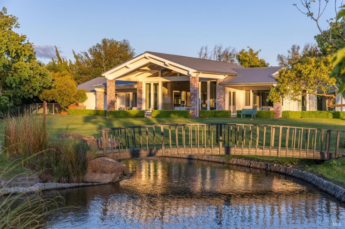  Modern single-story house with large windows and a covered patio, surrounded by greenery. A wooden bridge with railings crosses over a calm pond in the foreground. The house features brick pillars, a sloped roof, and a well-maintained lawn with hedges and trees.