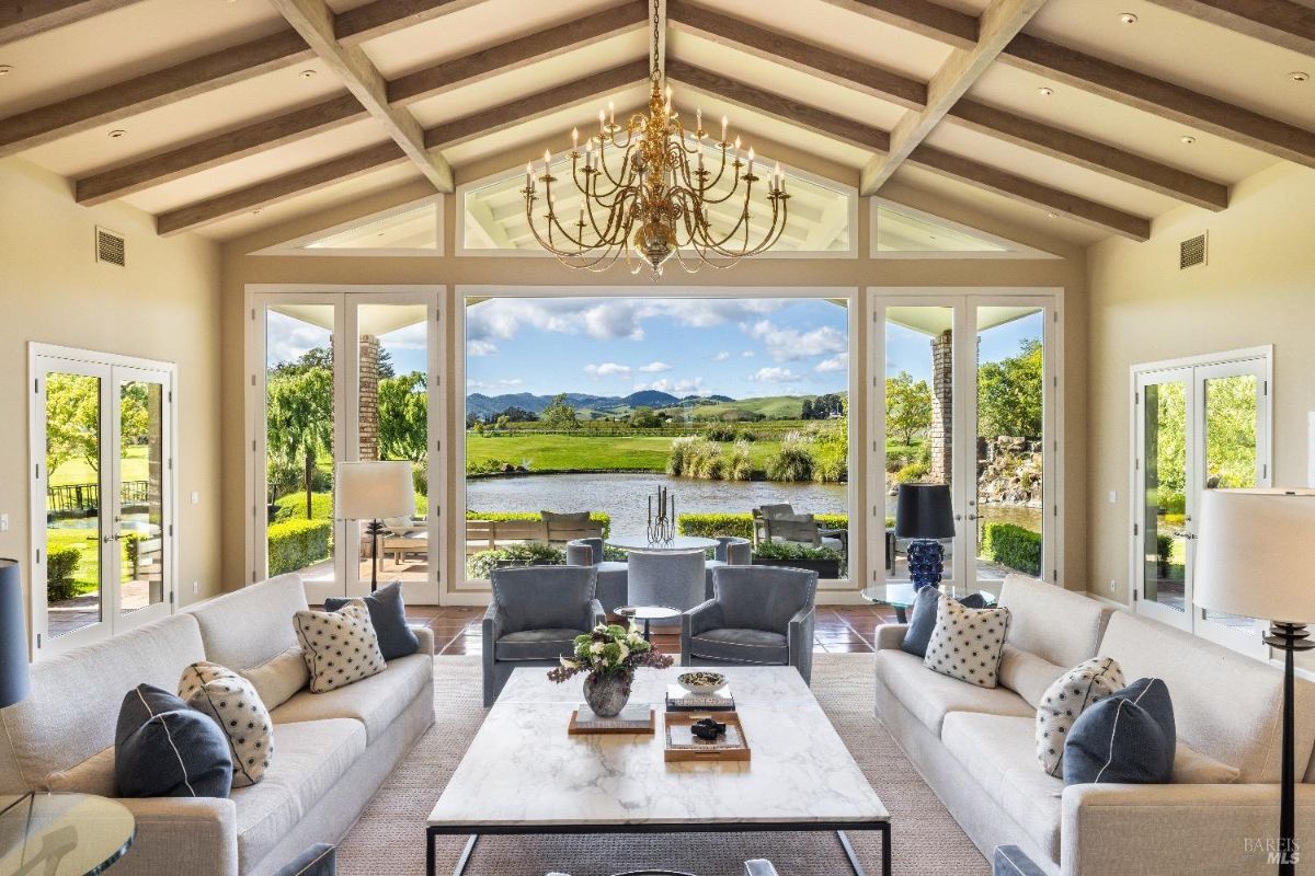 Spacious living room with large glass doors and windows offering a view of a pond and rolling hills. The room features two sofas and two armchairs, arranged around a marble coffee table, with decorative pillows and lamps. A chandelier hangs from the exposed wooden beam ceiling, and the outdoor area with patio furniture is visible through the doors.