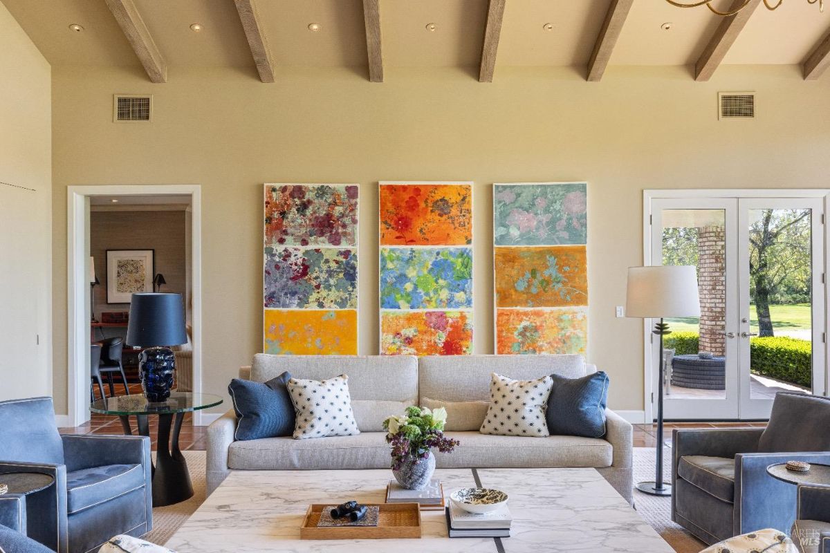 Living room with a central beige sofa, two armchairs, and a marble coffee table. Behind the sofa, there are three colorful abstract paintings mounted on the wall. To the sides of the sofa, there are two black lamps on small tables, with a doorway on the left and glass doors leading outside on the right.