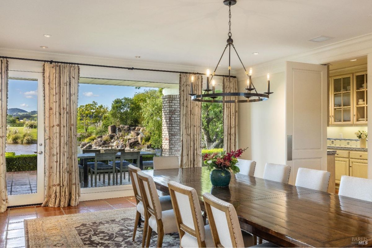  A dining room with a large wooden table surrounded by cushioned chairs. A chandelier with multiple exposed bulbs hangs above the table. Large glass doors and windows provide a view of the outdoor patio and natural landscape, including trees, a pond, and a rocky water feature.