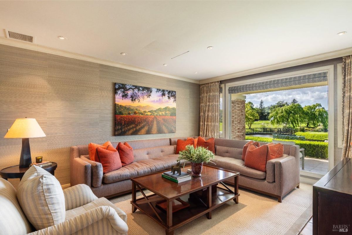 A living room with a large L-shaped sectional sofa adorned with orange throw pillows. A rectangular wooden coffee table with books and a decorative plant sits in front of the sofa. The room features a large window offering a view of a green outdoor landscape, while a painting of a vineyard hangs on the wall.