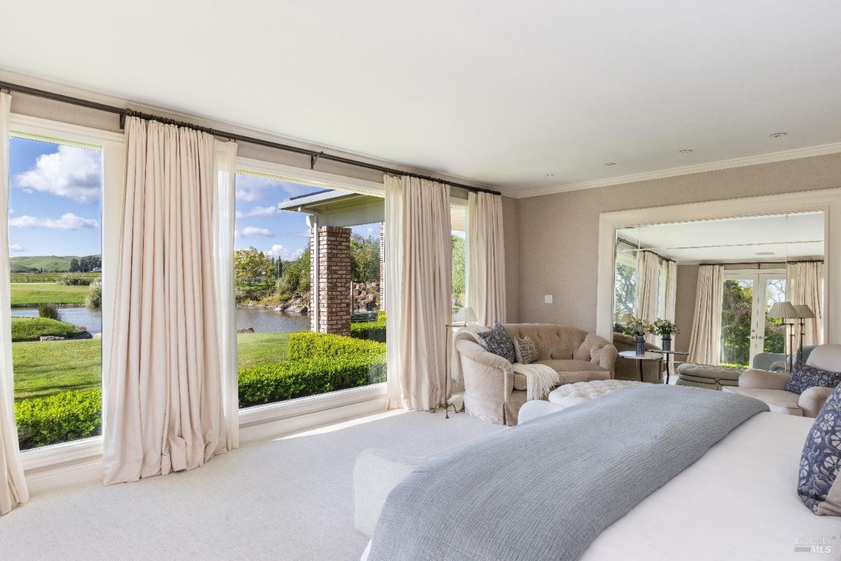 Bedroom with large windows covered by light-colored curtains, overlooking a green landscape with a pond. A seating area features a beige armchair, a matching loveseat, and decorative pillows. A large mirror reflects additional parts of the room, including more windows and a small table with a plant arrangement.