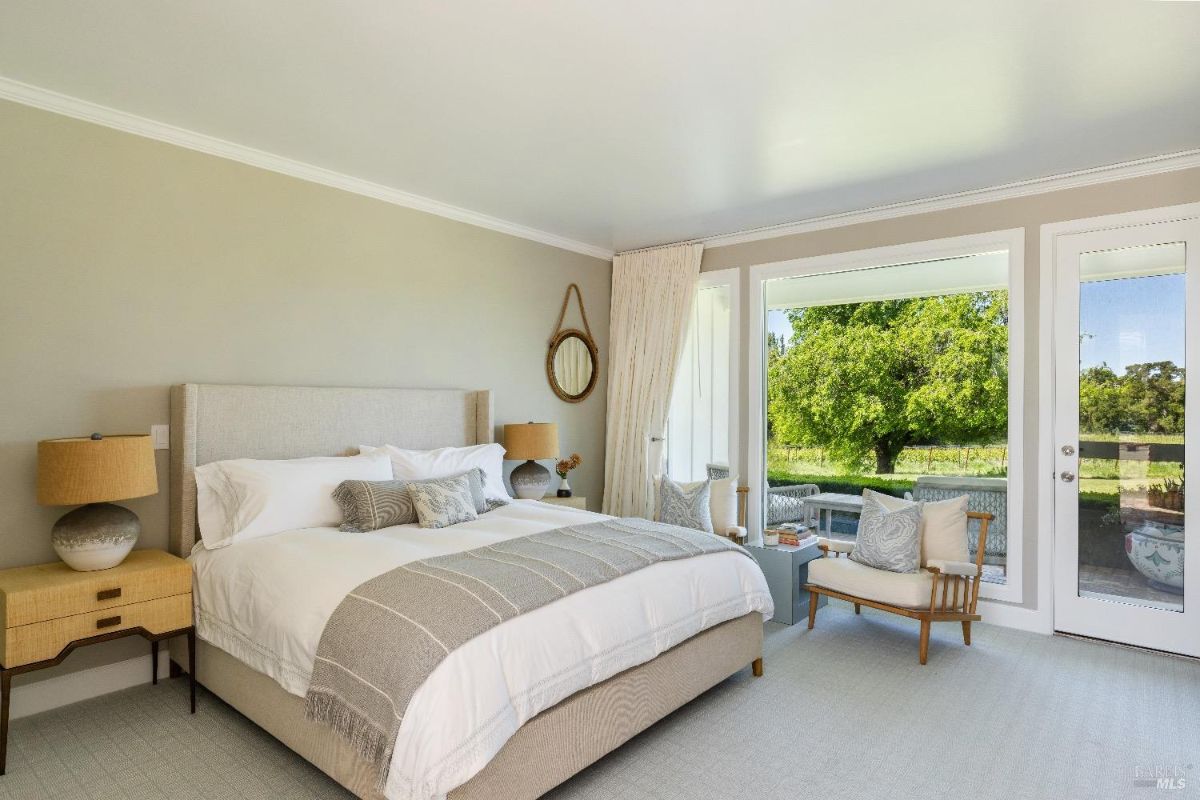 Bedroom with a large bed featuring a fabric headboard, white linens, and a decorative gray throw blanket. On either side of the bed are matching wooden nightstands with lamps, and there is a mirror mounted on the wall. The room has large windows and a glass door leading to an outdoor area with a view of green trees and a seating space outside.