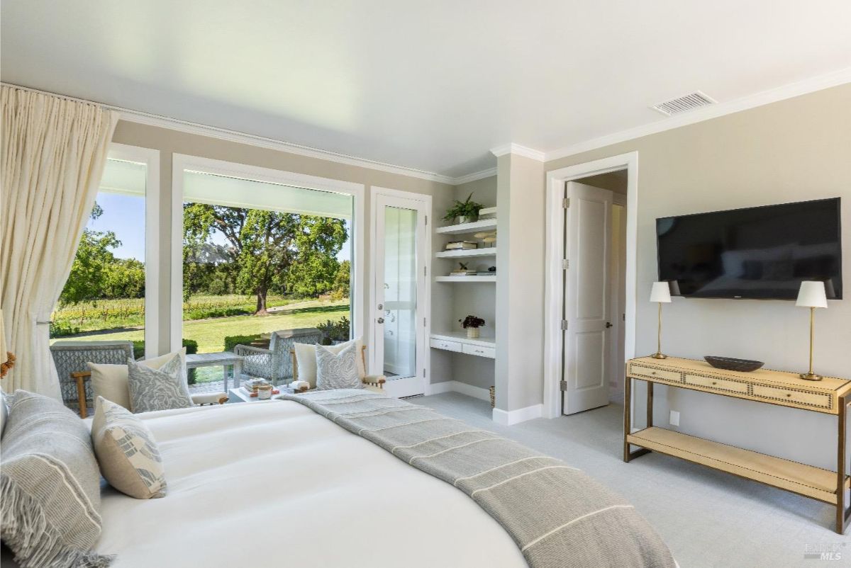 A large bed in the foreground, dressed in neutral bedding with decorative pillows. A built-in shelving unit with books and small plants is recessed into the wall near a doorway. A flat-screen TV is mounted on the wall above a console table, and a seating area outside the large windows overlooks a green outdoor landscape.