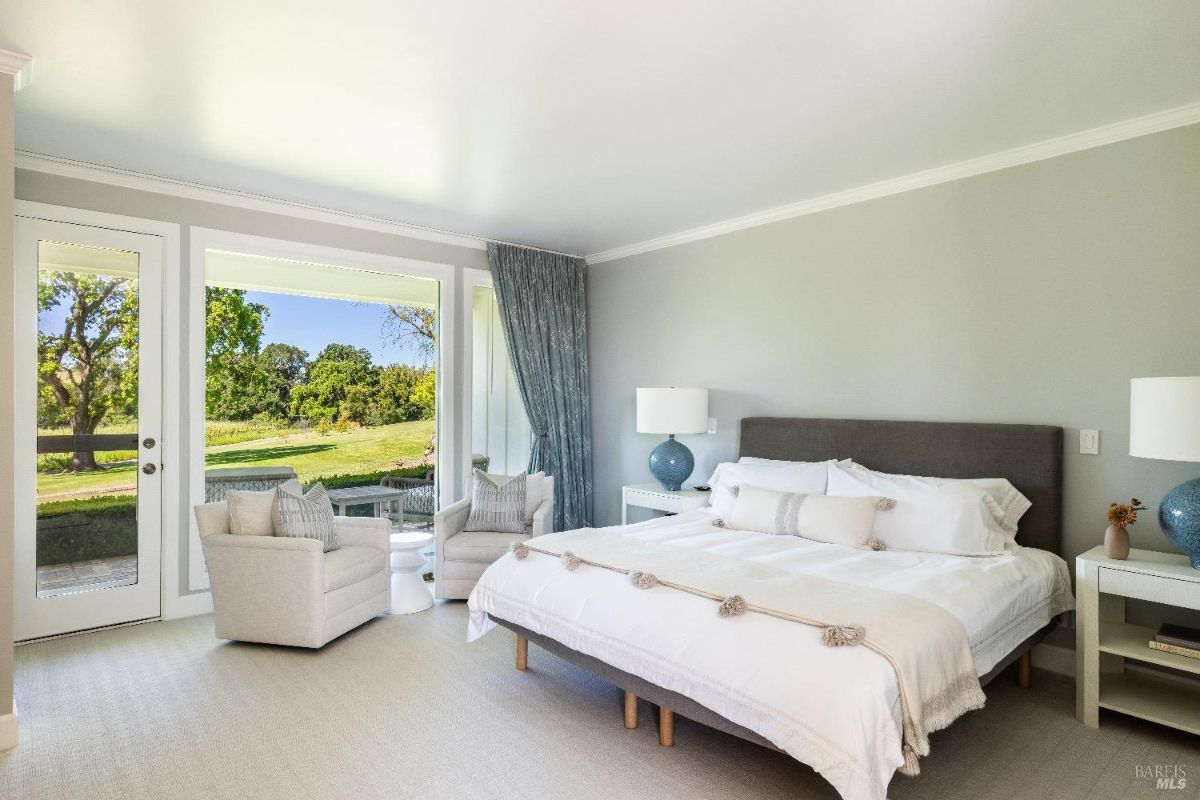 Bedroom with a large bed featuring a gray upholstered headboard, white bedding, and decorative pillows. Two matching blue table lamps sit on white nightstands on either side of the bed. The room has large windows and a glass door that provide a view of green trees and outdoor seating, with two armchairs and a small table placed near the window.