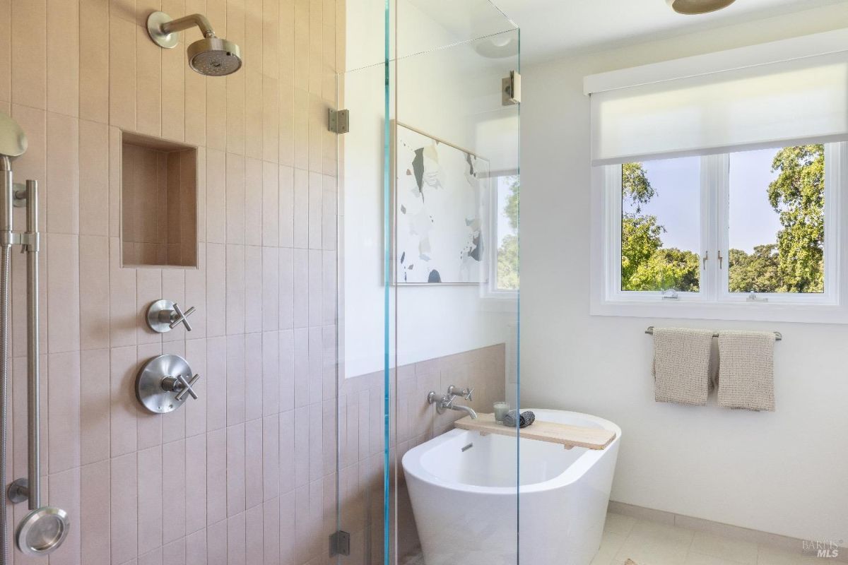 Bathroom with a glass-enclosed shower and a freestanding bathtub. The shower features a built-in niche, stainless steel fixtures, and tiled walls. A window with a roller shade allows natural light to enter, and two towels hang neatly on a bar below the window.