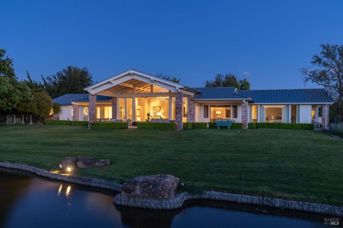 Modern single-story house with a large front porch and illuminated interior, surrounded by a green lawn at dusk. A pond with rocks borders the front of the lawn, reflecting the house lights. Trees and shrubs frame the house, and the sky above is transitioning into evening.