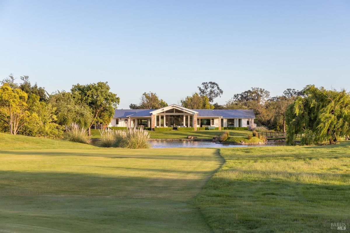 Modern house with a metal roof, surrounded by trees and greenery. A pond with tall grasses in the foreground reflects the house. The scene features a well-maintained lawn extending toward the house, with additional landscaping and a bridge visible near the water.