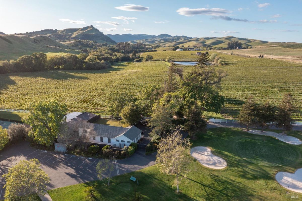 An aerial view of a rural landscape with a building situated near a vineyard. The building has a gray roof and is surrounded by trees, parking spaces, and open green areas. In the background, rolling hills, vineyards, and a pond are visible under a partly cloudy sky.